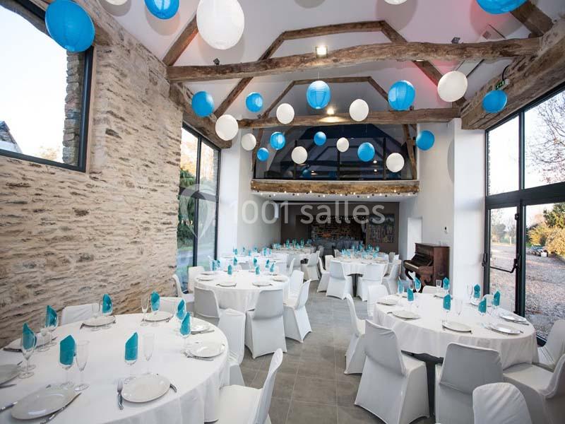 Location salle Plélan-le-Grand (Ille-et-Vilaine) - Domaine Le 5b #10 Salle de réception lumineuse avec tables rondes dressées, décorée de lanternes bleues et blanches suspendues au plafond.