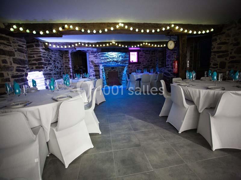 Location salle Plélan-le-Grand (Ille-et-Vilaine) - Domaine Le 5b #16 Salle en pierre avec tables rondes dressées, chaises blanches et guirlandes lumineuses suspendues au plafond.