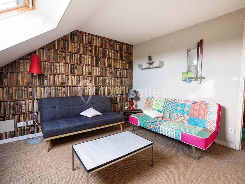 Location salle Plélan-le-Grand (Ille-et-Vilaine) - Domaine Le 5b #26 Salon mansardé avec canapé gris, canapé coloré, table basse en bois et mur tapissé d’un motif bibliothèque.