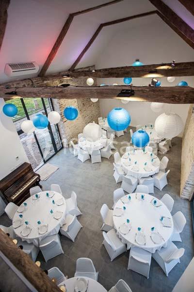 Location salle Plélan-le-Grand (Ille-et-Vilaine) - Domaine Le 5b #13 Salle de réception avec tables rondes dressées, décorée de lanternes bleues et blanches suspendues sous un plafond en…