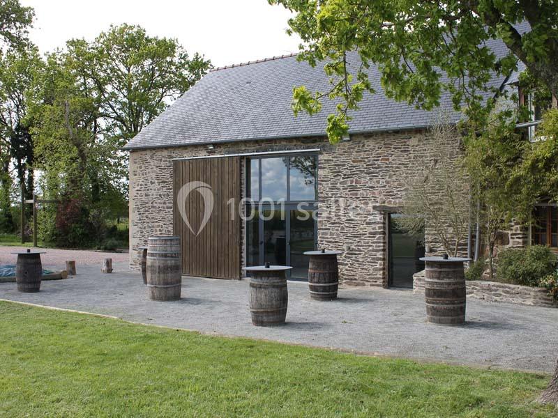 Location salle Plélan-le-Grand (Ille-et-Vilaine) - Domaine Le 5b #7 Bâtiment en pierre avec toit en ardoise, entouré de barriques en bois sur une cour gravillonnée et pelouse adjacente.