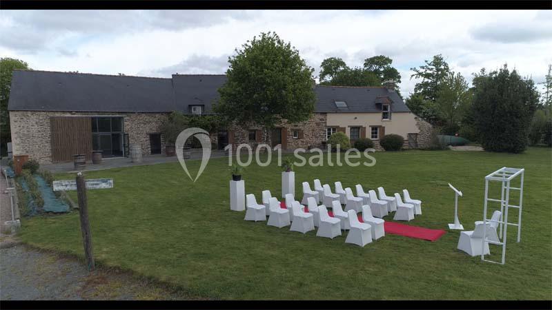 Location salle Plélan-le-Grand (Ille-et-Vilaine) - Domaine Le 5b #6 Disposition de chaises blanches sur une pelouse avec une arche et un tapis rouge devant un bâtiment en pierre.