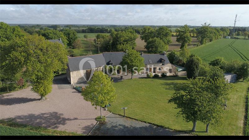 Location salle Plélan-le-Grand (Ille-et-Vilaine) - Domaine Le 5b #5 Vue aérienne d'une maison en pierre avec toit en ardoise, entourée d'un grand jardin et de champs verdoyants.