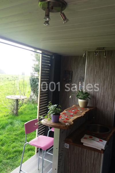 Location salle Plélan-le-Grand (Ille-et-Vilaine) - Domaine Le 5b #24 Vue d'un espace extérieur aménagé avec un comptoir en bois, des plantes en pot et des chaises roses près d'une pelouse.