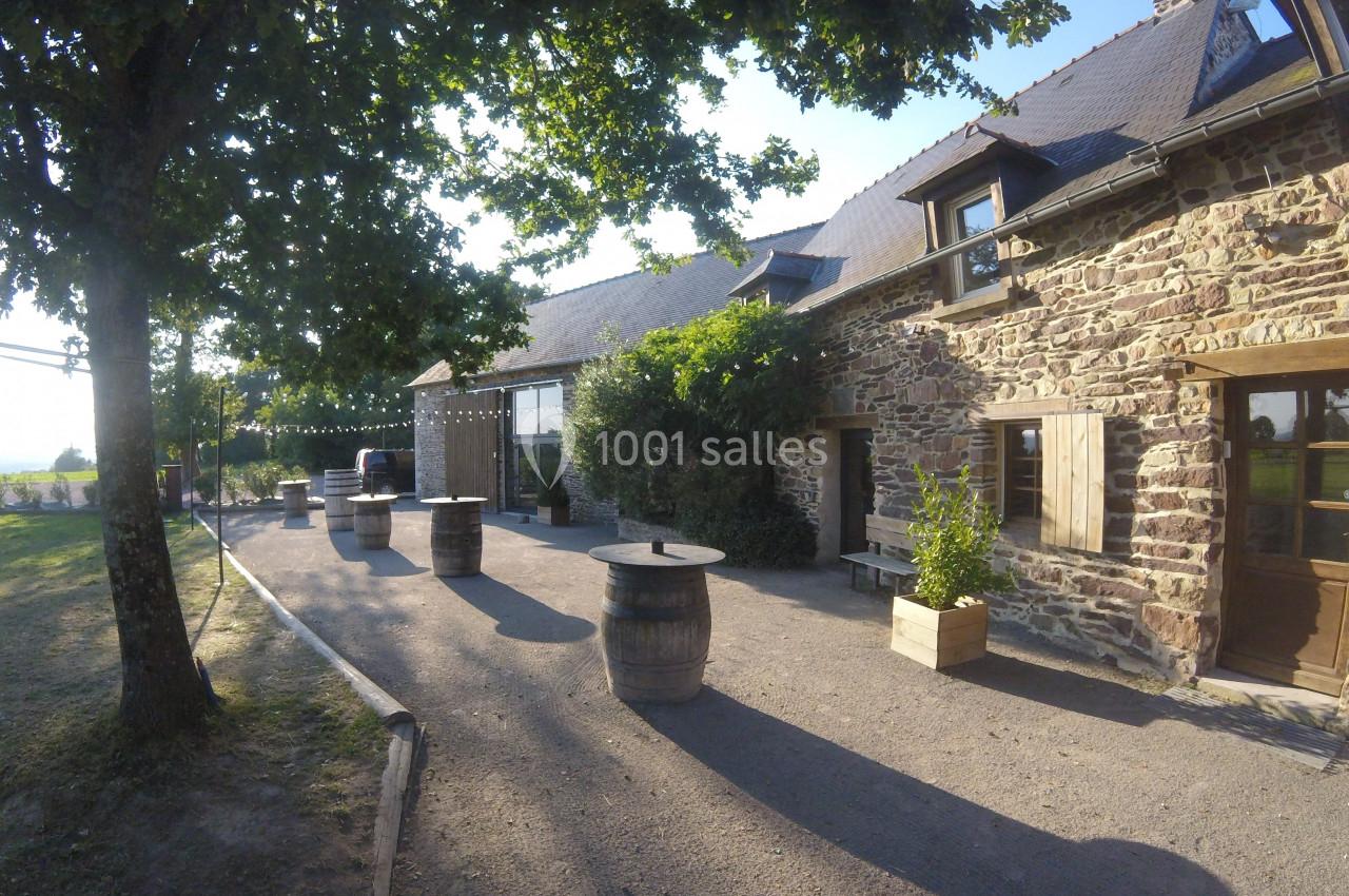 Location salle Plélan-le-Grand (Ille-et-Vilaine) - Domaine Le 5b #32 Cour extérieure d'une bâtisse en pierre avec barriques en guise de tables, entourée d'arbres et baignée de lumière.