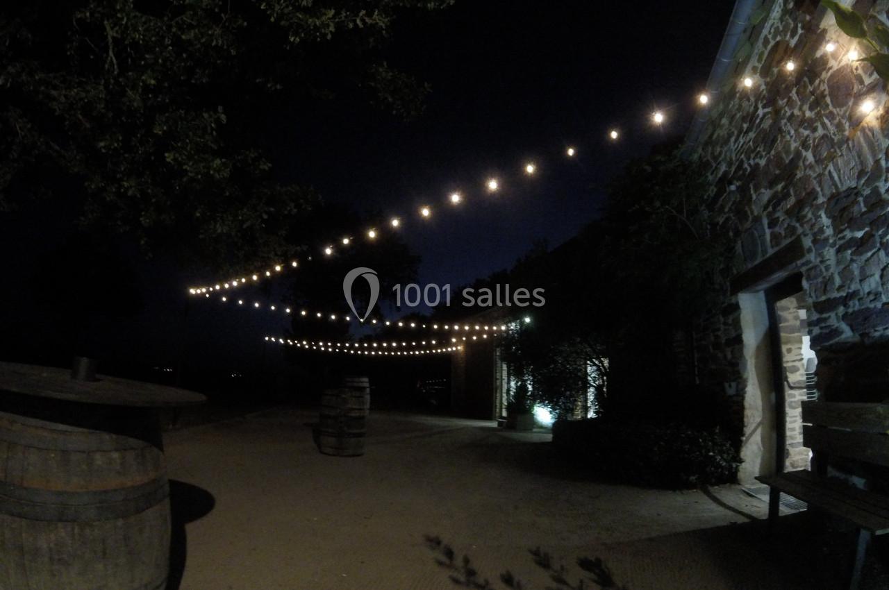 Location salle Plélan-le-Grand (Ille-et-Vilaine) - Domaine Le 5b #33 Guirlandes lumineuses suspendues au-dessus d'une cour extérieure sombre, bordée d'un bâtiment en pierre et de tonneaux.