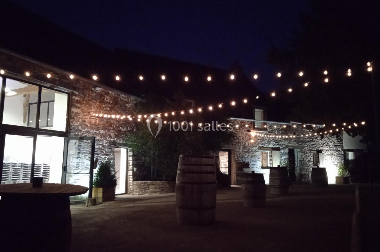 Location salle Plélan-le-Grand (Ille-et-Vilaine) - Domaine Le 5b #34 Cour extérieure en soirée avec guirlandes lumineuses suspendues, tonneaux en bois et bâtiments en pierre éclairés.