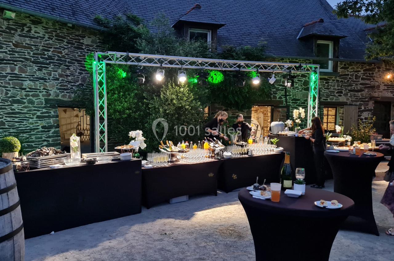 Location salle Plélan-le-Grand (Ille-et-Vilaine) - Domaine Le 5b #35 Buffet en extérieur avec tables hautes, éclairage nocturne et invités se servant dans un cadre en pierre.