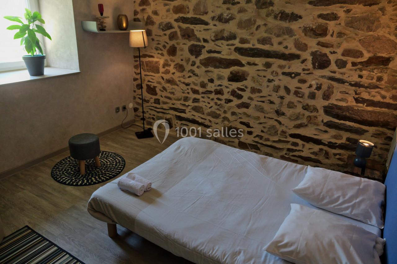 Location salle Plélan-le-Grand (Ille-et-Vilaine) - Domaine Le 5b #47 Lit bas avec draps blancs dans une chambre à mur en pierre, éclairée par une fenêtre et une lampe sur pied.