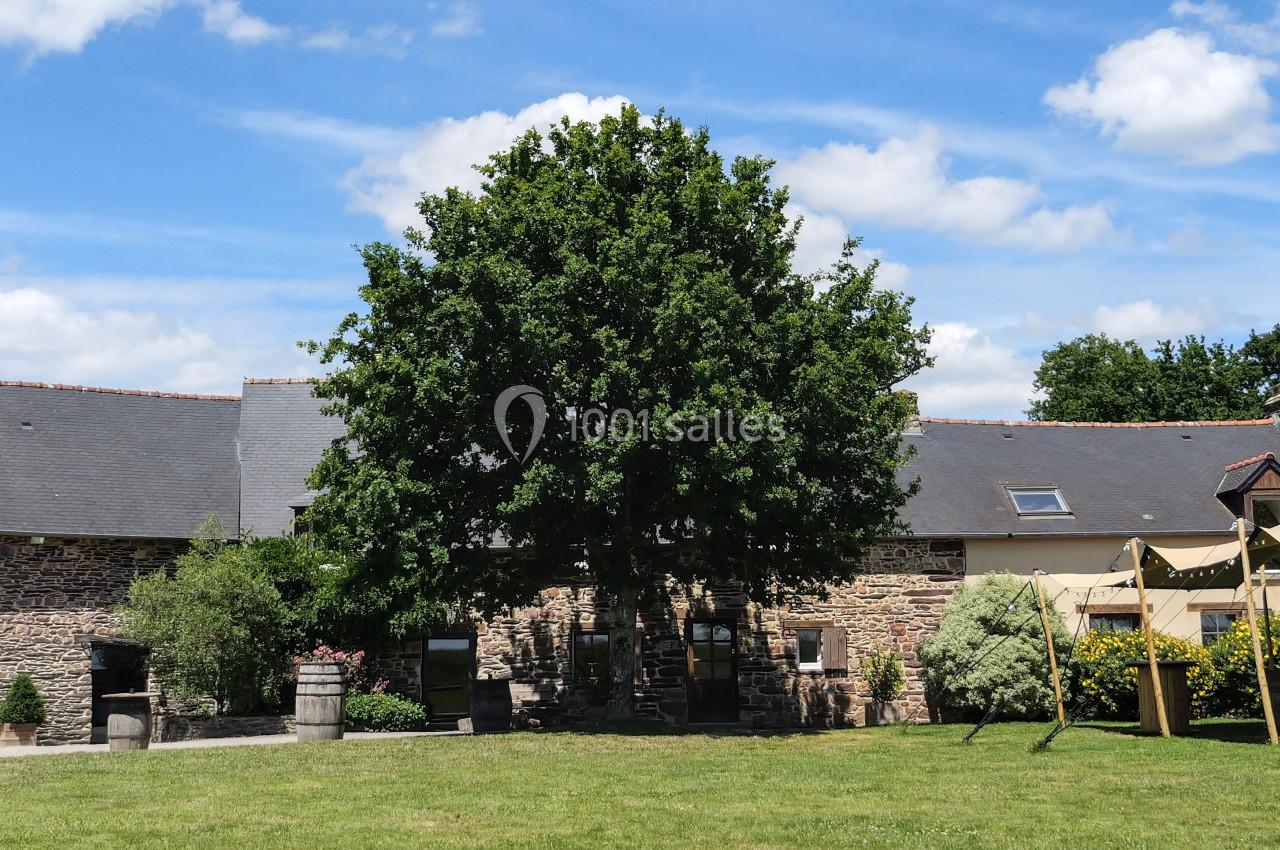 Location salle Plélan-le-Grand (Ille-et-Vilaine) - Domaine Le 5b #4 Bâtisse en pierre avec toit en ardoise, grand arbre central et tente beige sur une pelouse sous un ciel bleu.