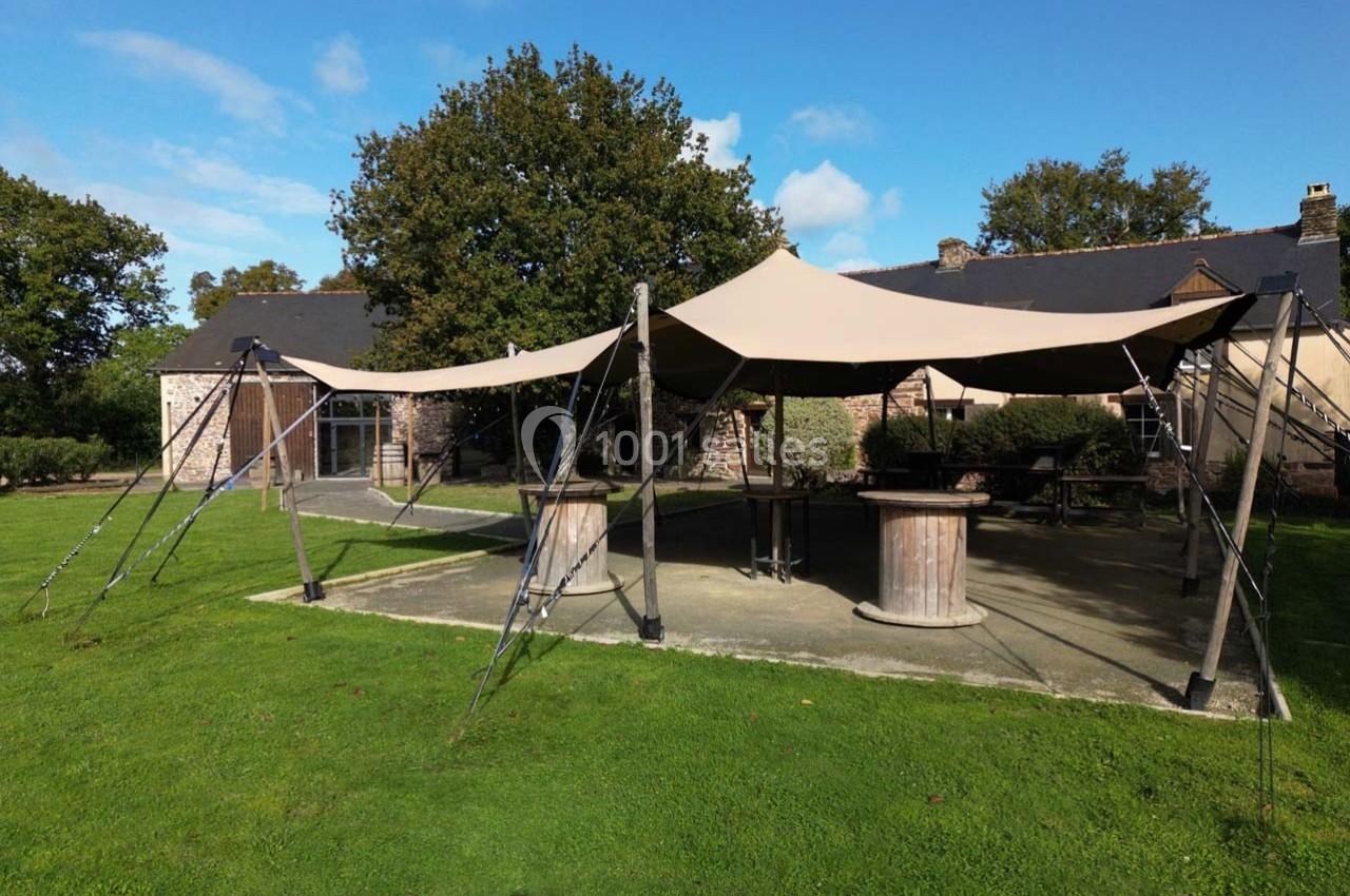 Location salle Plélan-le-Grand (Ille-et-Vilaine) - Domaine Le 5b #3 Tente beige montée sur une terrasse en pierre, entourée de verdure et d'arbres, devant un bâtiment en pierre.