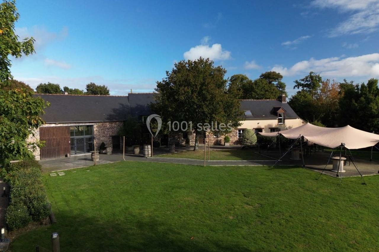 Location salle Plélan-le-Grand (Ille-et-Vilaine) - Domaine Le 5b #2 Bâtiment en pierre avec toit en ardoise, entouré d'un grand jardin verdoyant et d'une tente beige installée à droite.