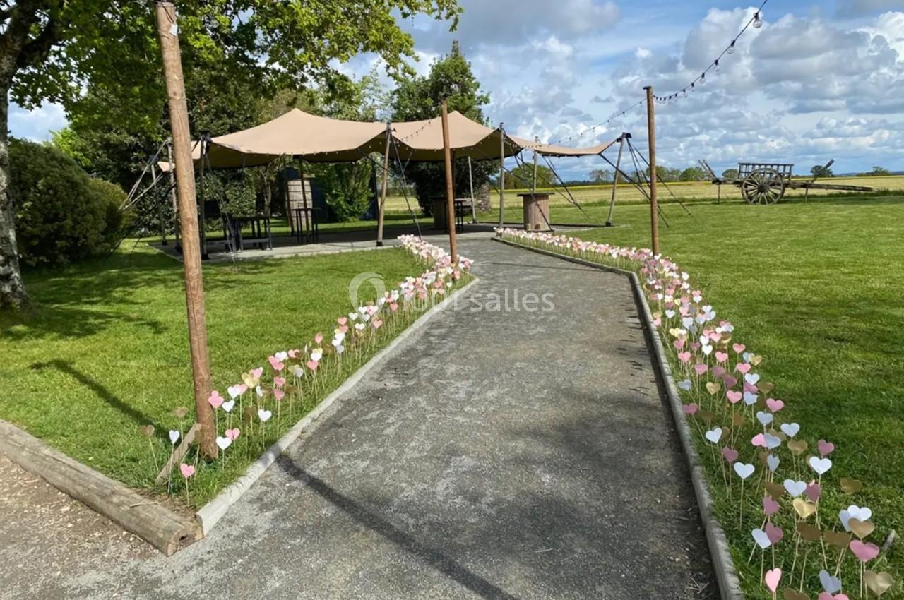 Location salle Plélan-le-Grand (Ille-et-Vilaine) - Domaine Le 5b #48 Allée bordée de petits cœurs en papier menant à des tentes beige dans un espace vert sous un ciel partiellement nuageux.