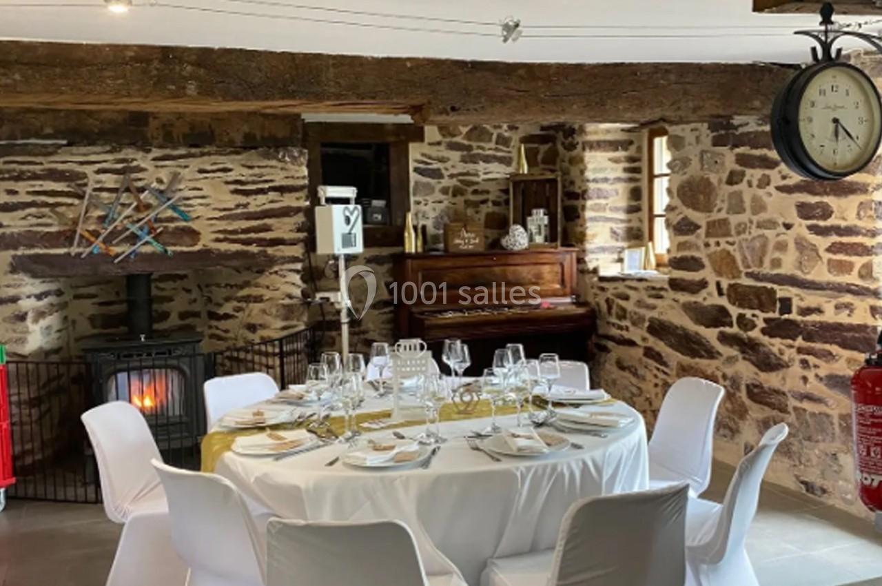 Location salle Plélan-le-Grand (Ille-et-Vilaine) - Domaine Le 5b #50 Salle rustique avec murs en pierre, table ronde dressée pour un repas, poêle allumé et piano en arrière-plan.