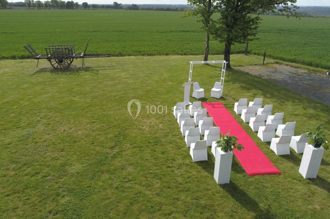 Location salle Plélan-le-Grand (Ille-et-Vilaine) - Domaine Le 5b #54 Chaises blanches disposées en extérieur autour d'une allée rouge, avec un arche décoratif et un champ en arrière-plan.