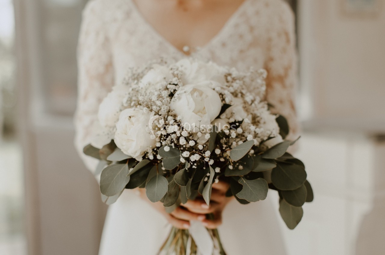 Bouquet de mariée composé de roses blanches, de gypsophiles et de feuillage, tenu par une personne en robe claire.