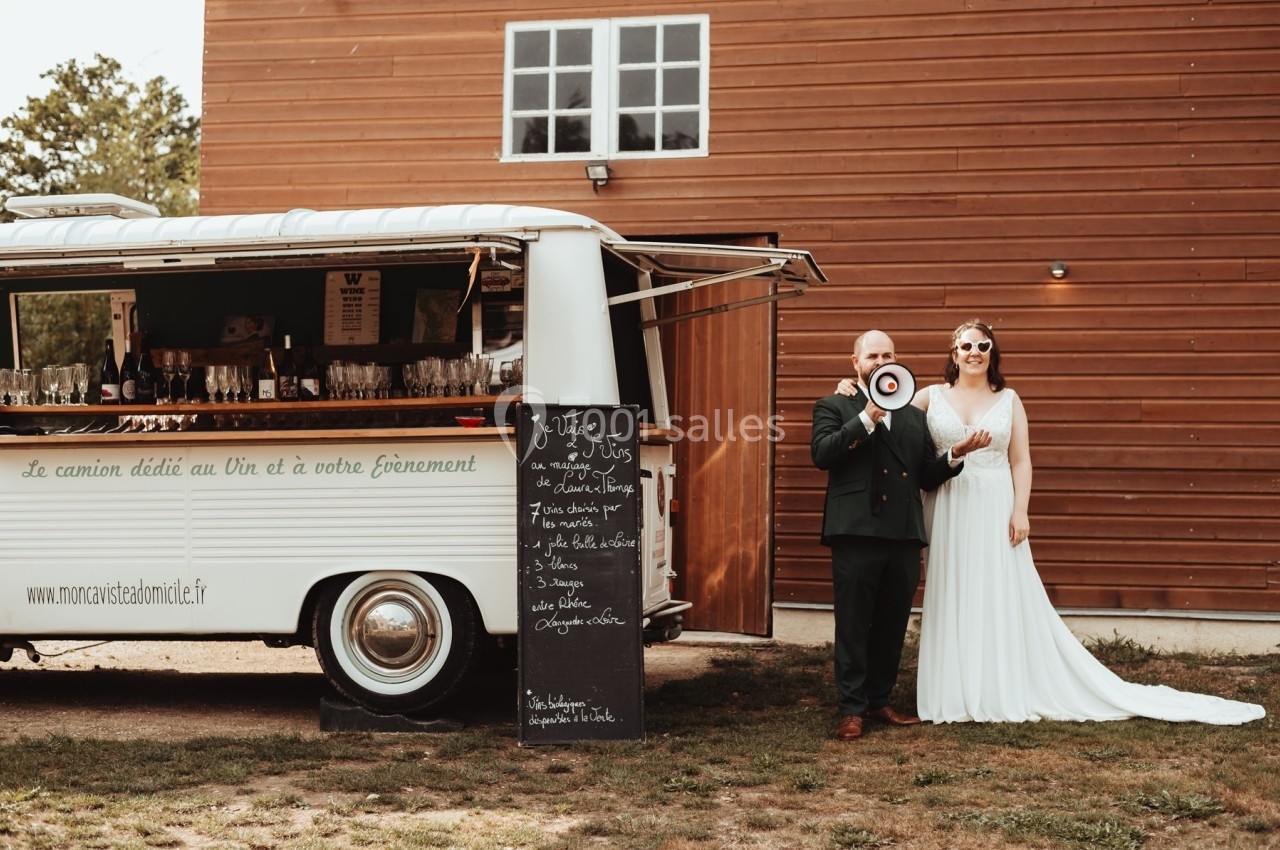 Un couple en tenue de mariage pose devant un camion-bar vintage blanc, avec une ardoise et des verres exposés.