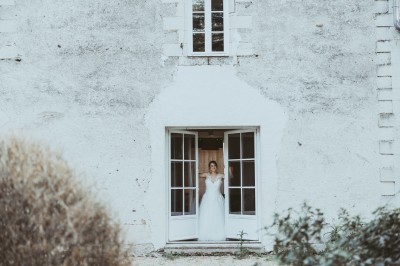 Robe de mariée en dentelle suspendue devant une fenêtre lumineuse avec vue sur un jardin verdoyant.