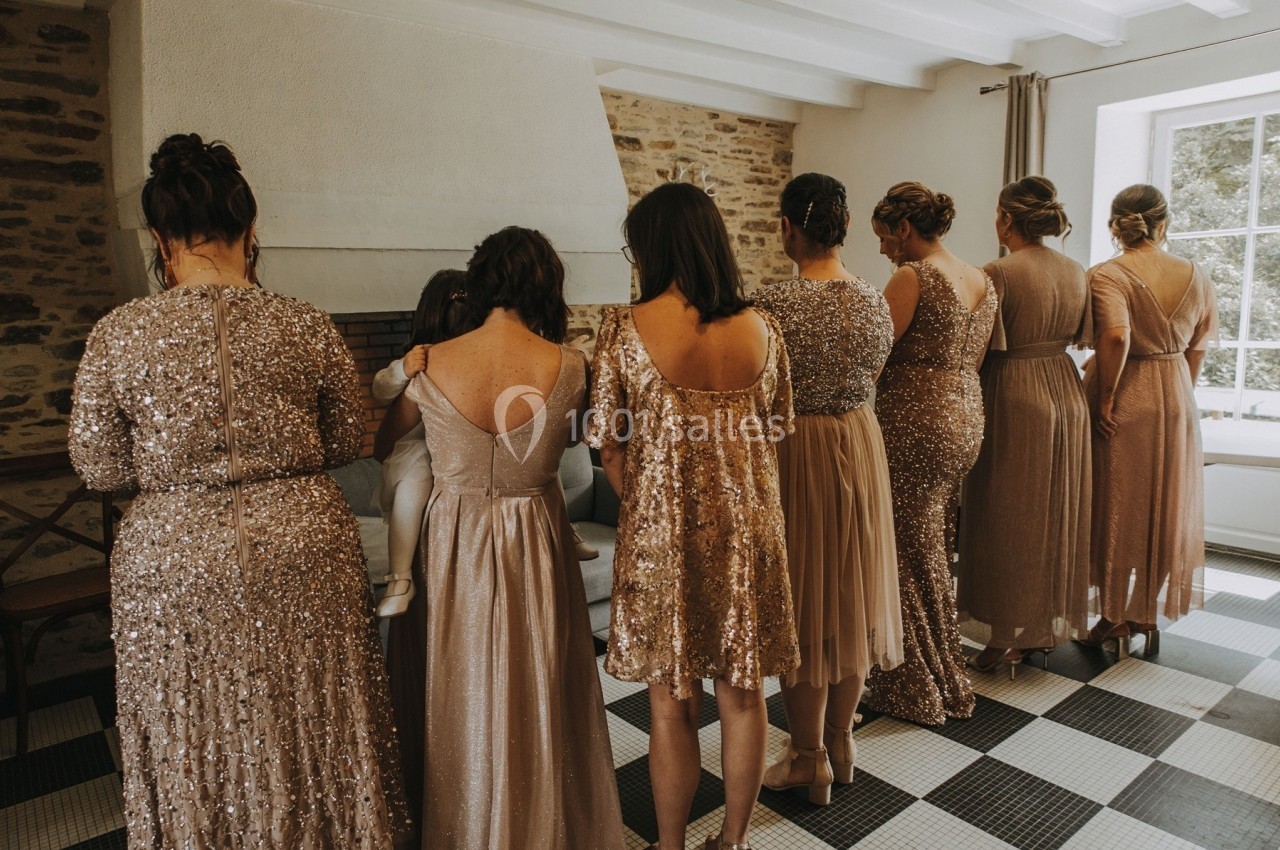 Des femmes en robes scintillantes, vues de dos, se tiennent dans une pièce lumineuse avec un mur en pierre apparente.