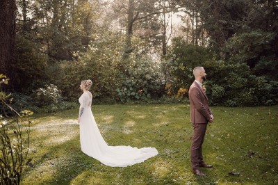 Robe de mariée en dentelle suspendue devant une fenêtre lumineuse avec vue sur un jardin verdoyant.