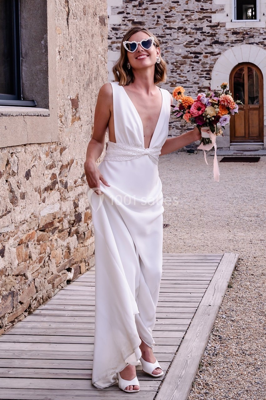 Femme souriante en robe de mariée blanche tenant un bouquet de fleurs colorées devant un bâtiment en pierre.