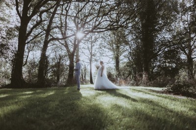 Robe de mariée en dentelle suspendue devant une fenêtre lumineuse avec vue sur un jardin verdoyant.