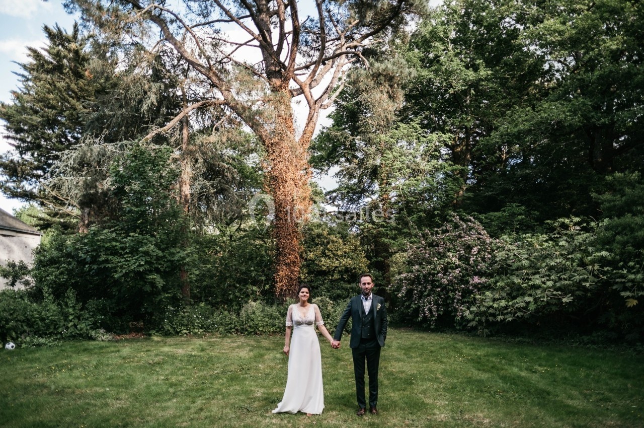 Un couple en tenue de mariage se tient la main dans un jardin verdoyant, entouré d'arbres et de buissons.