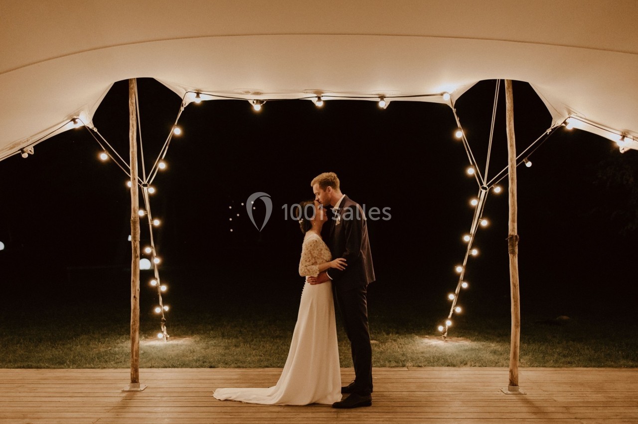 Un couple se tient sous une tente éclairée par des guirlandes lumineuses, dans un cadre nocturne.