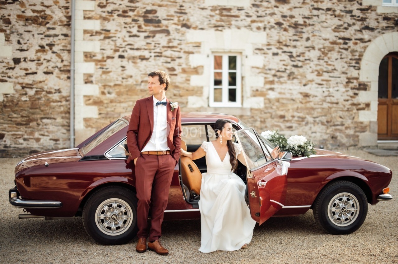 Un couple en tenue de mariage pose près d'une voiture ancienne rouge devant un bâtiment en pierre.