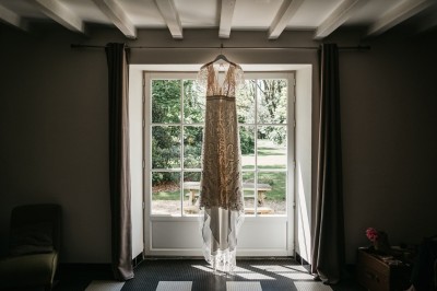 Robe de mariée en dentelle suspendue devant une fenêtre lumineuse avec vue sur un jardin verdoyant.