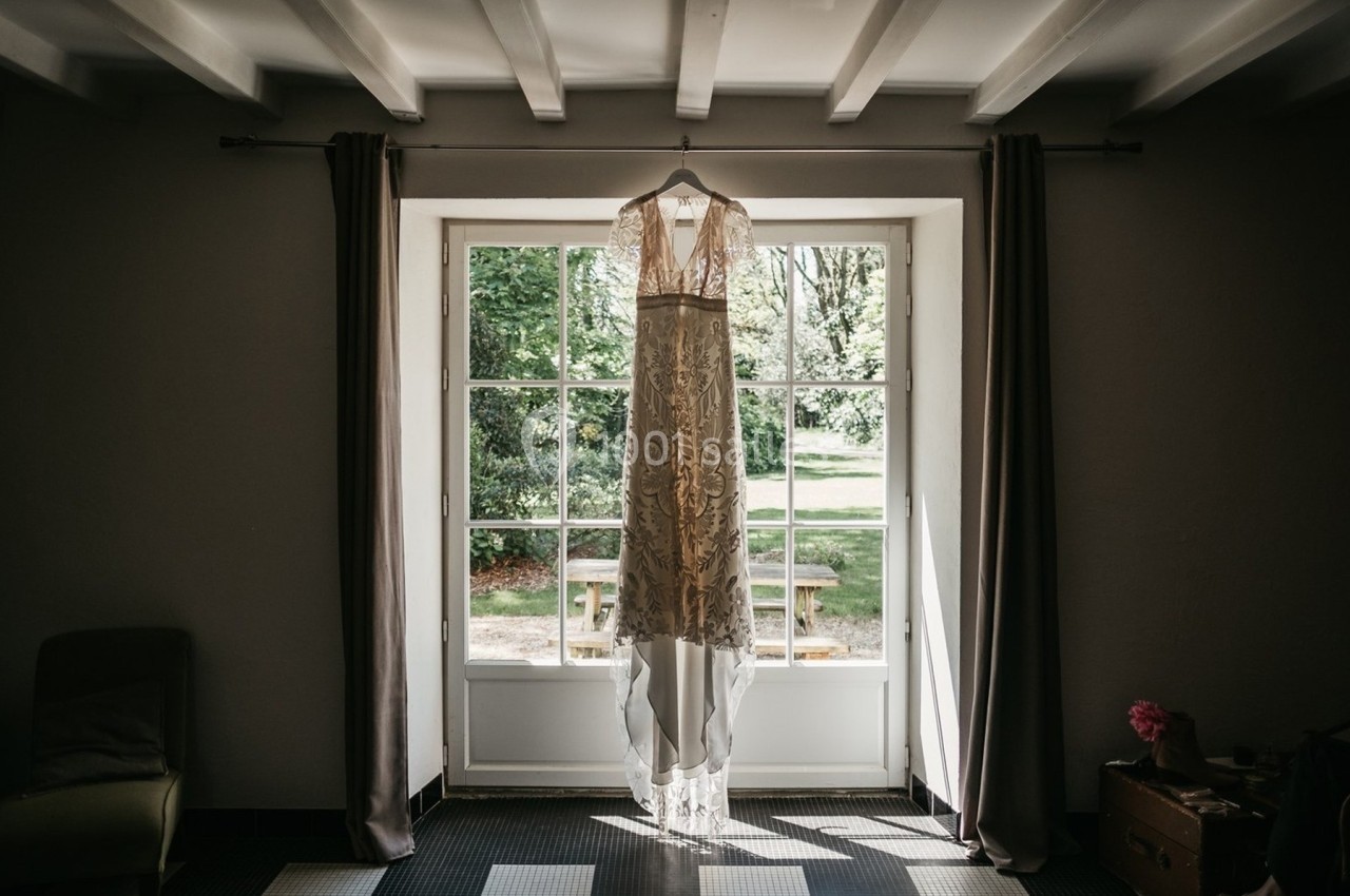 Robe de mariée en dentelle suspendue devant une fenêtre lumineuse avec vue sur un jardin verdoyant.