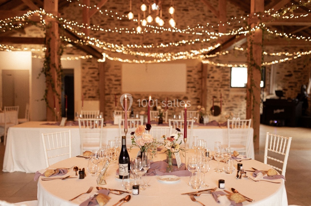 Salle de réception décorée avec des guirlandes lumineuses, tables dressées avec nappes blanches et centre de table floral.