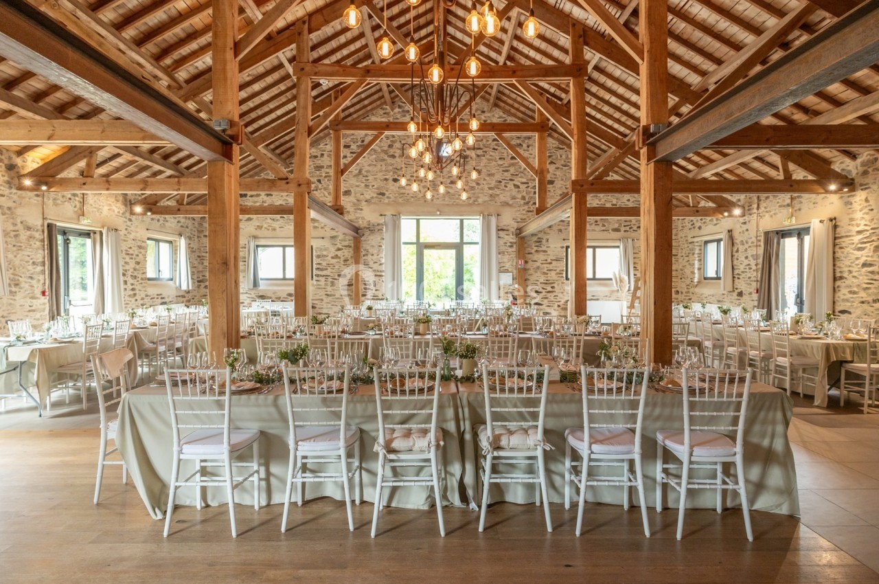 Salle de réception lumineuse avec charpente en bois, tables décorées et chaises blanches alignées.
