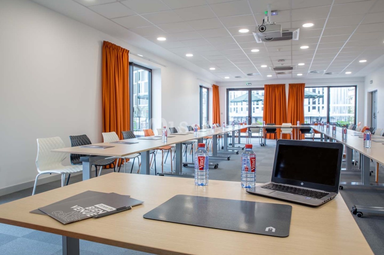Salle de réunion lumineuse avec tables disposées en U, chaises modernes, rideaux orange et équipement de projection.