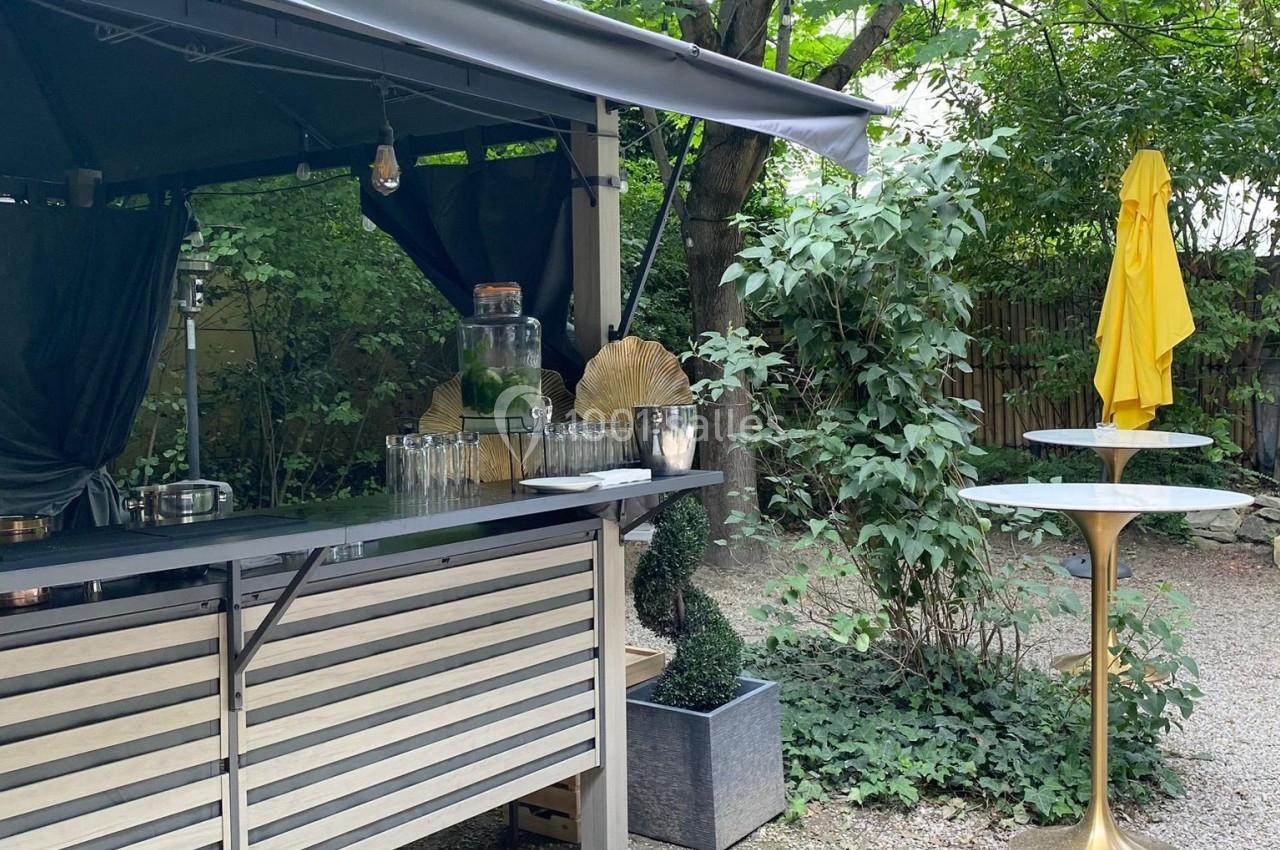 Stand extérieur avec comptoir en bois et métal, entouré de végétation, avec une table haute et un parasol jaune.
