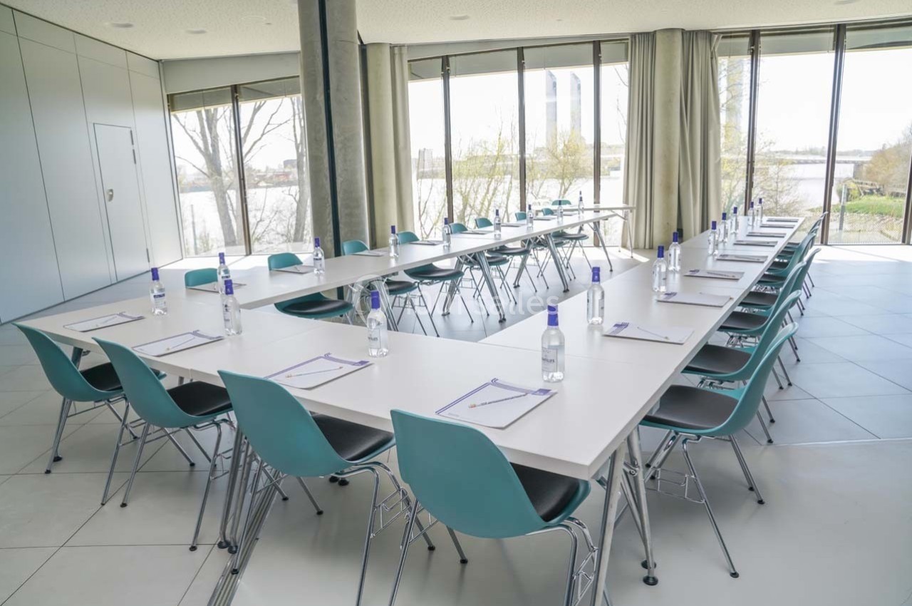 Salle de réunion lumineuse avec tables disposées en U, chaises turquoise et fournitures de bureau sur chaque place.