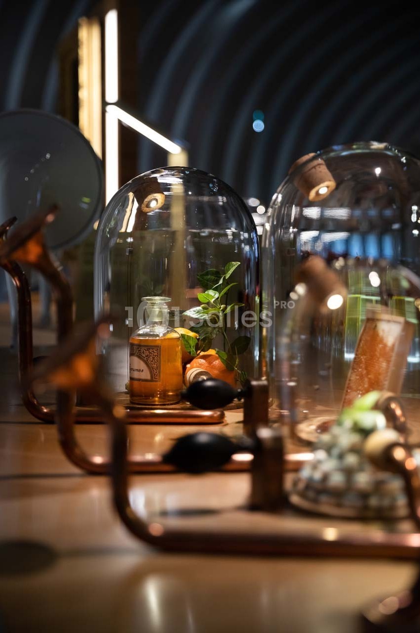 Flacons et éléments décoratifs sous cloches en verre sur une table éclairée dans un espace moderne et tamisé.