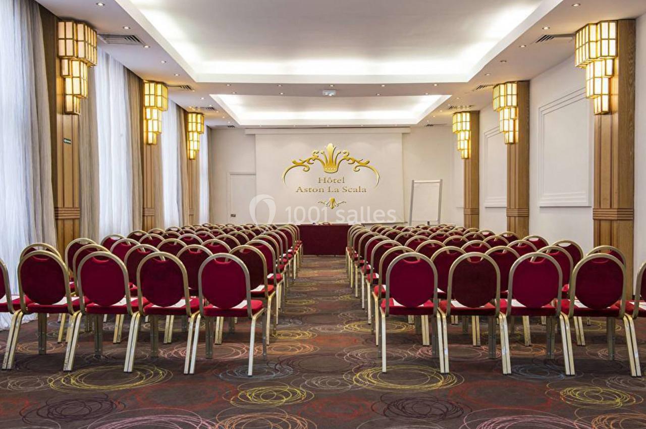 Salle de conférence lumineuse avec rangées de chaises rouges alignées face à un mur décoré et un tableau blanc.