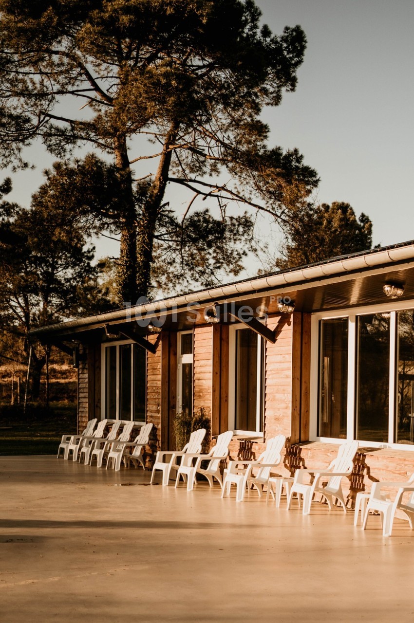 Façade d'un bâtiment en bois avec de grandes fenêtres, entouré de chaises blanches et d'arbres sous une lumière douce.