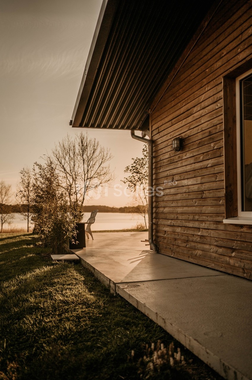 Façade en bois d'une maison avec terrasse, vue sur un lac au coucher du soleil et arbres en arrière-plan.