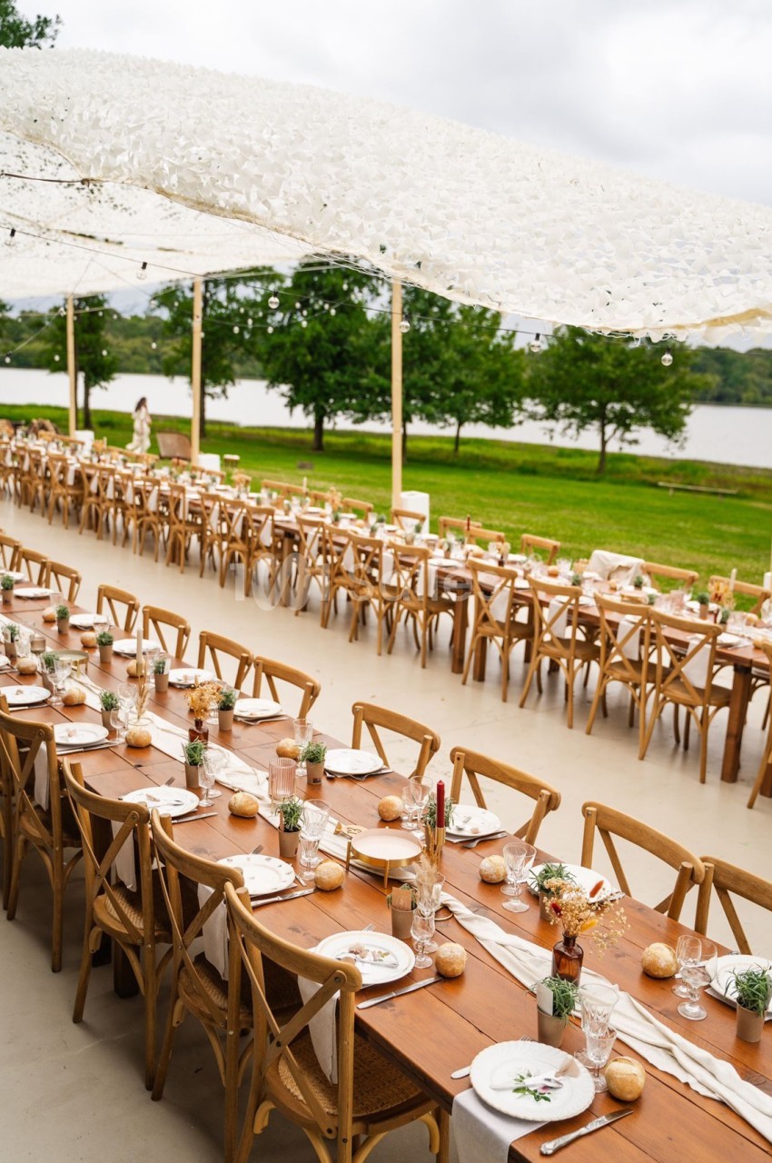 Tables en bois décorées pour un repas en plein air sous une toile blanche, avec vue sur un lac et des arbres.