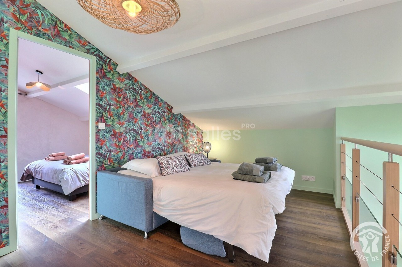 Chambre mansardée avec lit double, canapé, mur à motifs floraux et vue sur une seconde chambre en arrière-plan.