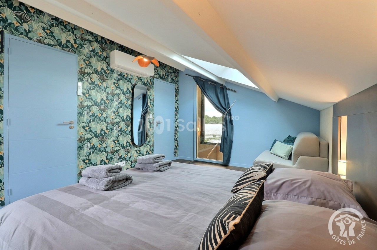 Chambre mansardée avec lit double, décoration murale végétale, fauteuil, fenêtre et serviettes pliées sur le lit.