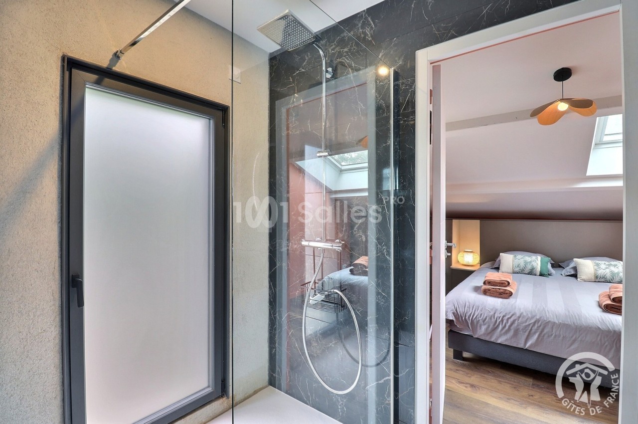 Salle de bain moderne avec douche à l'italienne, vue sur une chambre lumineuse avec lit double et décoration soignée.