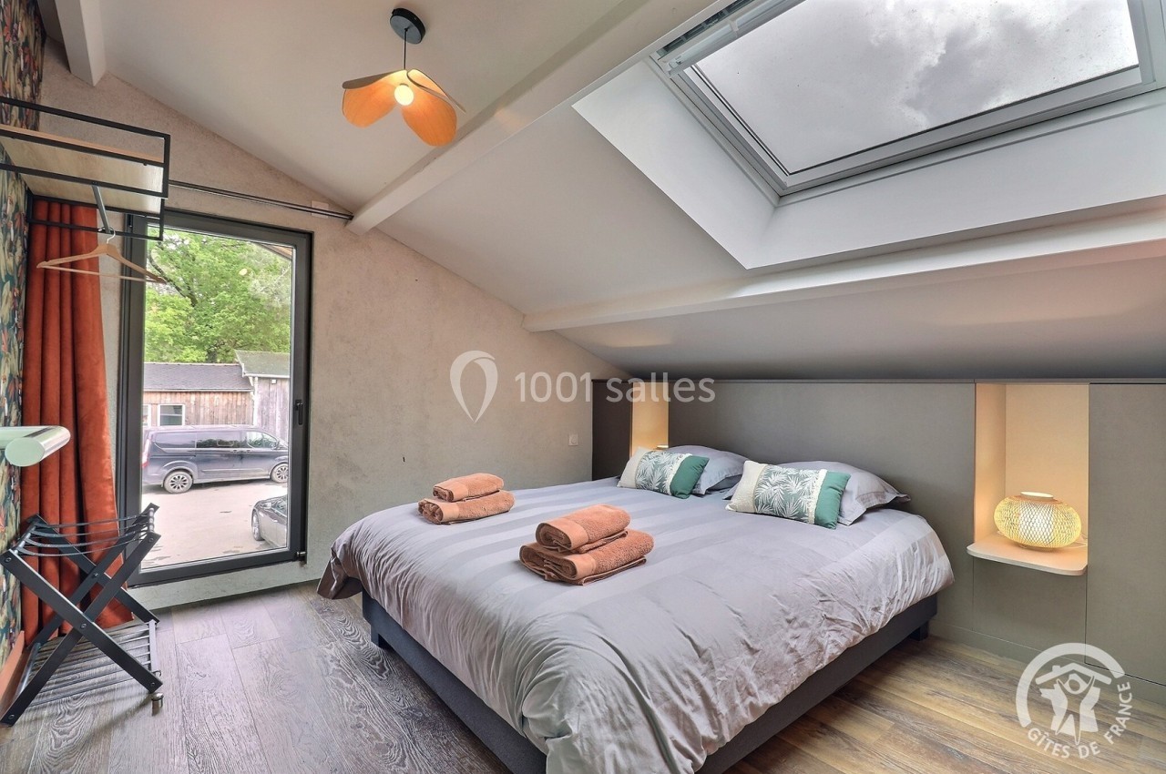 Chambre lumineuse avec lit double, linge de lit plié, grande fenêtre, velux et vue sur l'extérieur verdoyant.