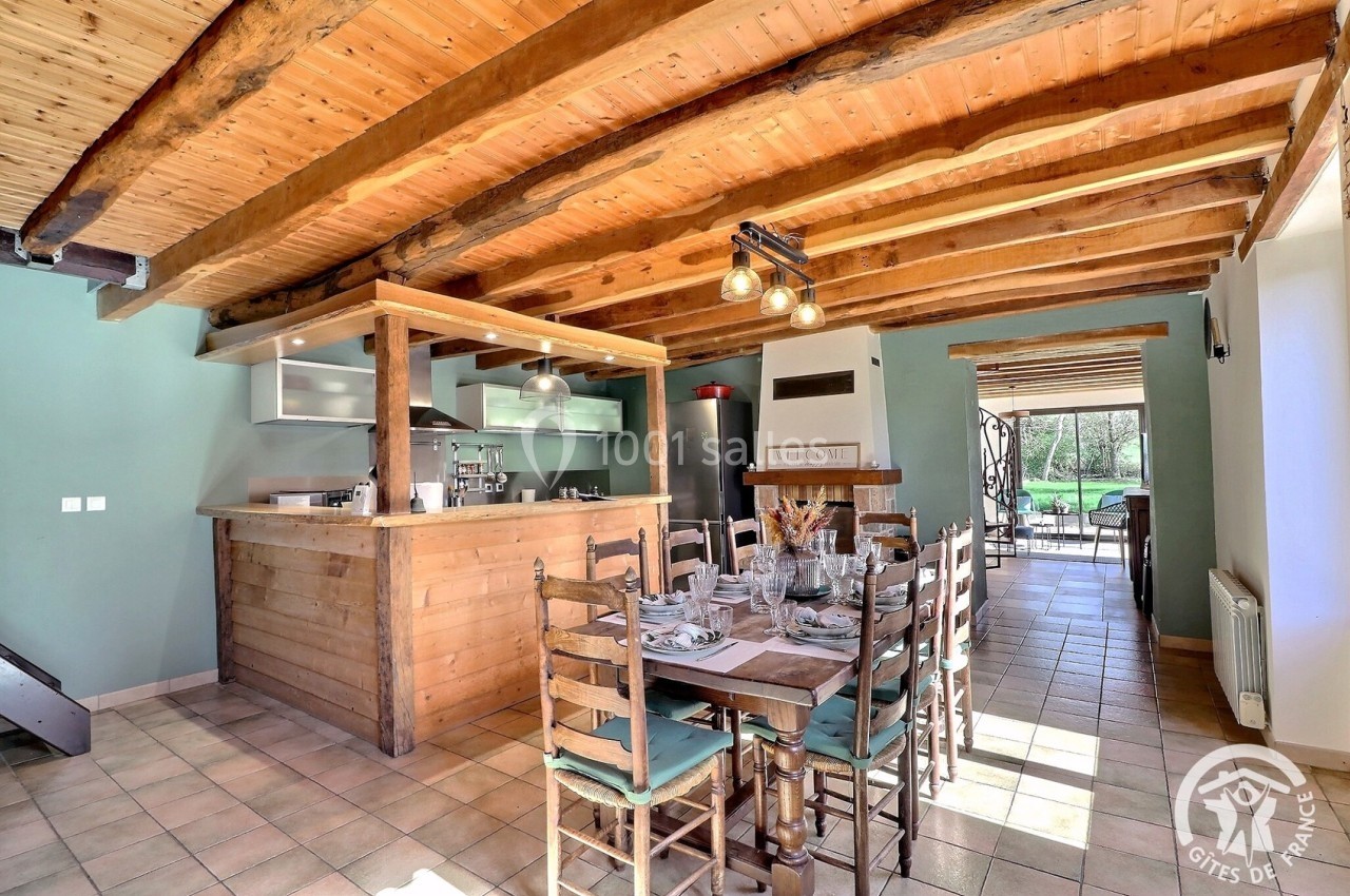 Salle à manger lumineuse avec table en bois, cuisine ouverte et poutres apparentes, donnant sur un jardin.