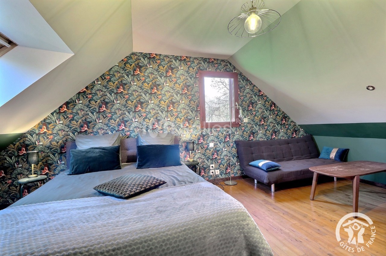 Chambre mansardée avec lit double, canapé, table basse et mur décoré d'un papier peint à motifs tropicaux.