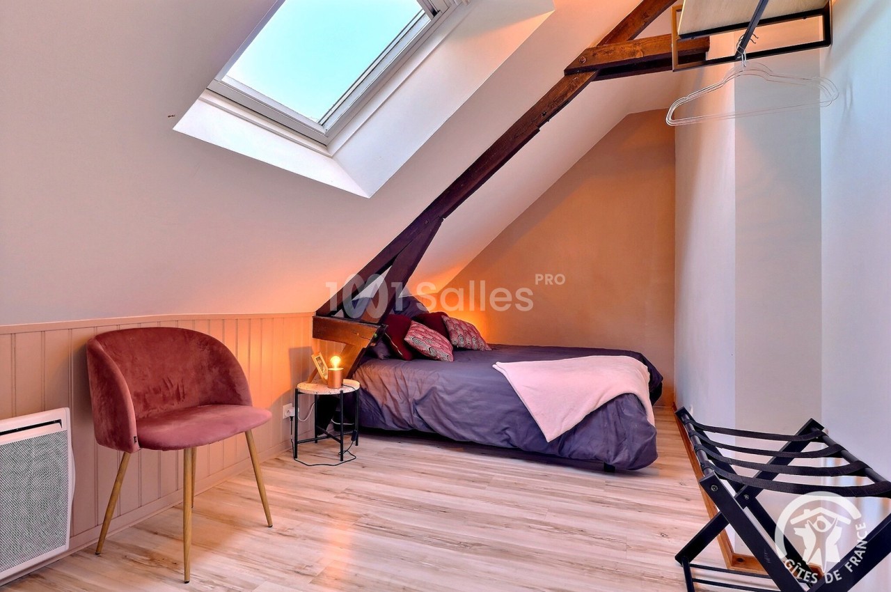 Chambre sous les combles avec lit double, fauteuil rose, poutres apparentes et lumière naturelle d'une fenêtre de toit.