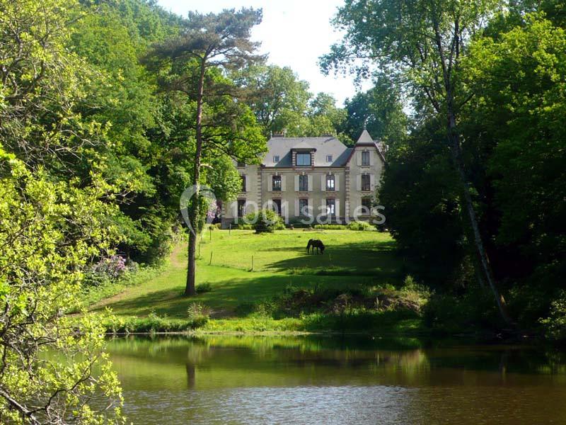 Manoir en pierre entouré de verdure, avec un cheval sur une pelouse en pente, près d'un étang calme.