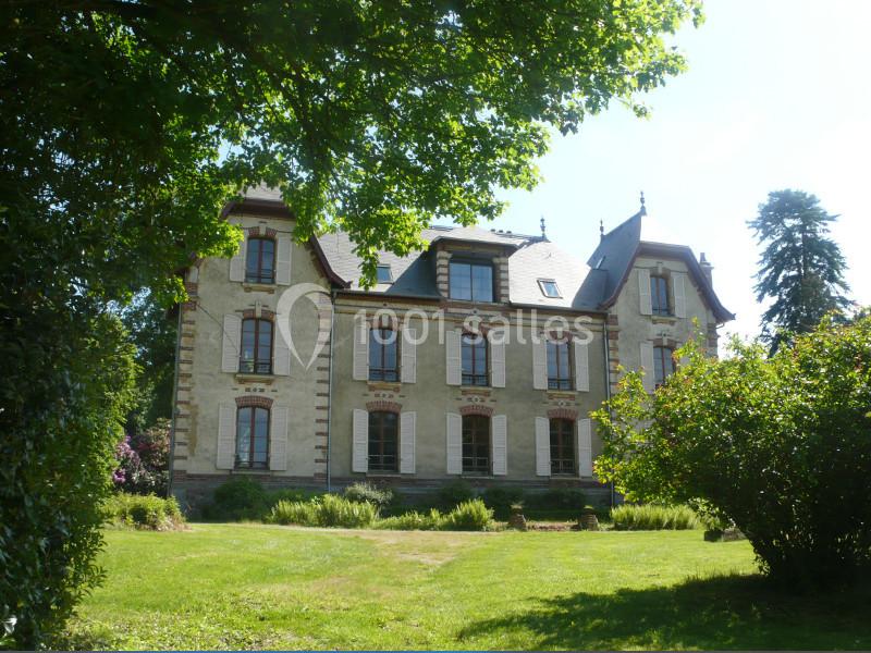 Grande maison en pierre avec volets blancs, entourée d'arbres et d'un jardin verdoyant sous un ciel clair.
